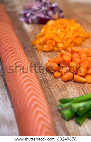 Fresh raw salmon on a wooden chopping board in a domestic kitchen, next to spring onions, chopped carrots, yellow pepper, and red onion. Photo has short depth of field with focus on the carrots.
