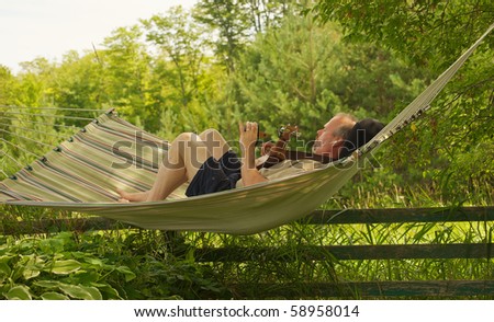 A middle aged man composing music with a guitar and hand held recording device in a hammock in the shade in the summer.