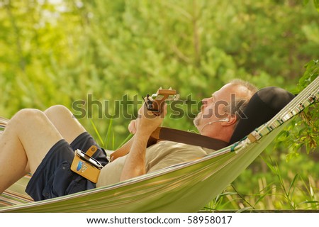 A middle aged man composing music with a guitar and hand held recording device in a hammock in the shade in the summer.