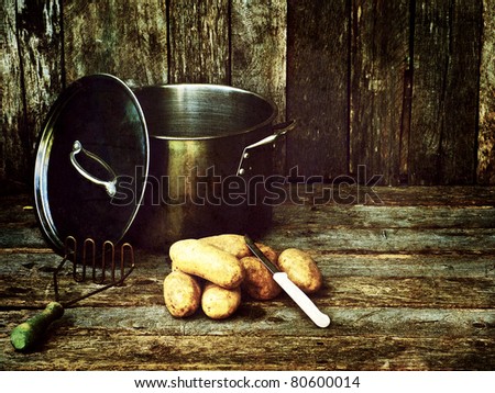 Antique textured, richly colored moody vintage style image of potatoes, a knife, old fashioned potato masher and pot on grunge wooden backdrop with copy space.