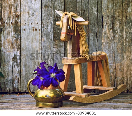 Old rocking horse  with purple clematis flowers in an antique copper vase on a rustic wood backdrop.