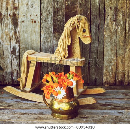 Old rocking horse  with orange lilies and daisies in an antique copper vase on a rustic wood backdrop.