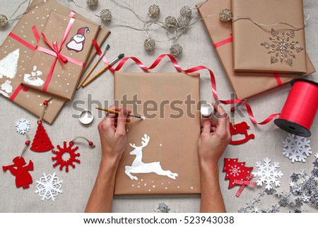 Hands decorating wrapped christmas gifts. Drawing a deer with white paint on brown craft paper of parcel. Top view of present decor accessories.