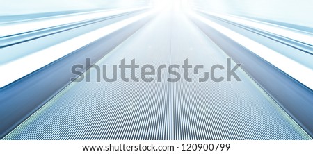 Perspective wide angle view of modern light blue illuminated and spacious high-speed moving commercial escalator with fast blurred trail of handrail in vanishing traffic motion in airport corridor