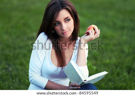 Pretty girl-student laying on the lawn, reading a textbook and eating apple
