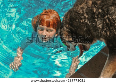 red haired child swimming in pool with her pet dog looking down from the edge at her.