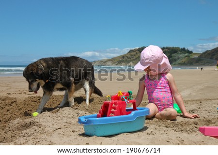 little red headed girl in pink bathing suit on beach making sand castles with her pet dog digging at its ball nearby