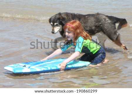 little red headed girl in wetsuit running into sea carrying surf board with black and white pet dog following.