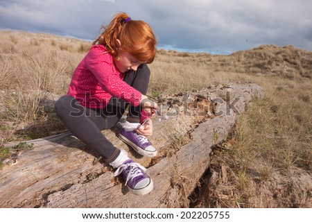 little red haired girl tying up her shoelaces while sitting on a log at the beach