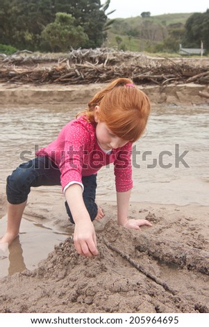 A little red haired girl building a sand castle witn wet sand beside a tidal river lagoon at a New Zealand surf beach