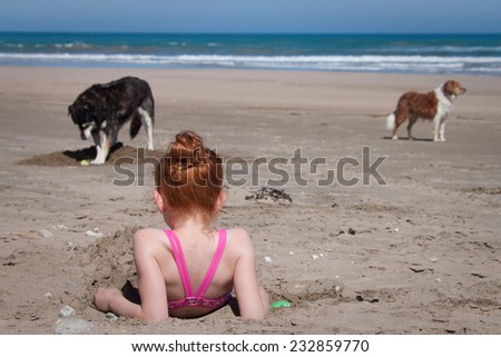 little red haired girl lying in sand at beach watching her dogs play