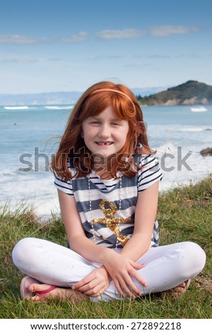 little red haired girl wearing nautical t shirt sitting on a grassy bank above a surf beach