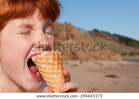 close up of young red haired girl eating an ice cream biting waffle cone, eyes shut, at a beach in New Zealand