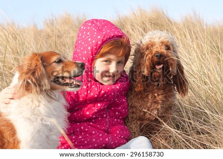 red haired girl in red winter jacket hugging two pet dogs - a red haired collie type dog and a brown curly coated breed on a cold winter\'s day in grassy sand dunes at a beach in Gisborne, New Zealand