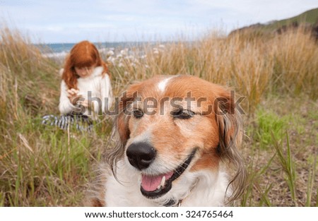 little red haired girl with red haired collie type dog sitting among long dune grass on a sand dune at a surf beach, near Gisborne, East Coast, North Island, New Zealand