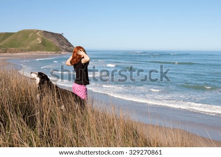 red haired girl and her pet black dog looking out over picturesque New Zealand surf beach from high sand hills above