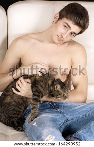 Beautiful young man with naked torso sits on a white leather couch and holds a cat