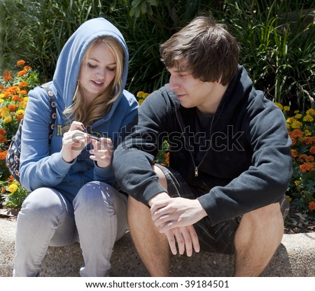 Teenage boy and girl, 18-years-old, sitting and talking on a curb with flowers behind them.  Both are wearing sweatshirts and are looking at something the girl is holding.