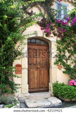 Richly-grained wooden door to private stucco villa in medieval village of Mougins, France.  Flowering vines frame the doorway, and there\'s a classic blue-shuttered window above.