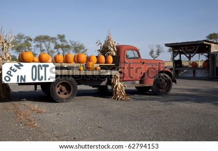 Pumpkins on an old red farm truck with a sign advertising that the farm stand will be open in October.