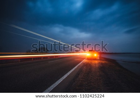 The car stopped at the roadside by night. Evening traffic of a country road. Red lights. Light headlights in the form of lines. Blue toning.