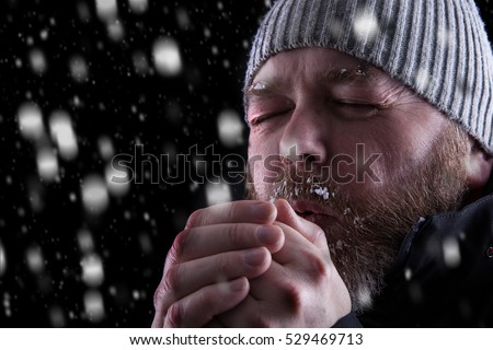 Freezing cold man standing in a snow storm blizzard trying to keep warm. Eyes closed and blowing warm air into his hands. Wearing a hat and coat with frost and ice on his beard and eyebrows.
