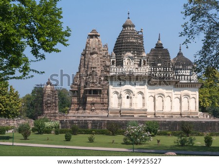 Old and younger Hindu temple, built by Chandela Rajputs, at Western site in India\'s Khajuraho framed by trees. White grey for the younger and older beige structure against blue skies over green grass.