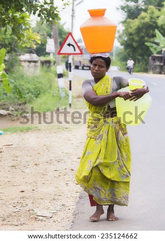 KUMBAKONAM, INDIA - OCTOBER 10, 2013: Along the road, a woman in yellow-green sari carries an orange and a yellow water jar, one on her head, one on her hip.