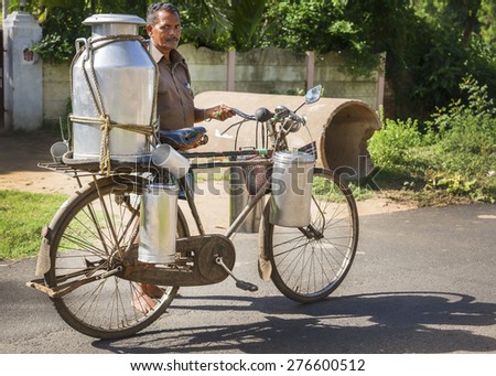 KUMBAKONAM, INDIA - OCTOBER 13, 2013:  Milkman carrying a very large and small milk cans on his rusty bike along a rural road.