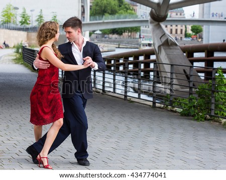 two young people - a man in black suit and a woman wearing red dress - dancing tango outside under the bridge