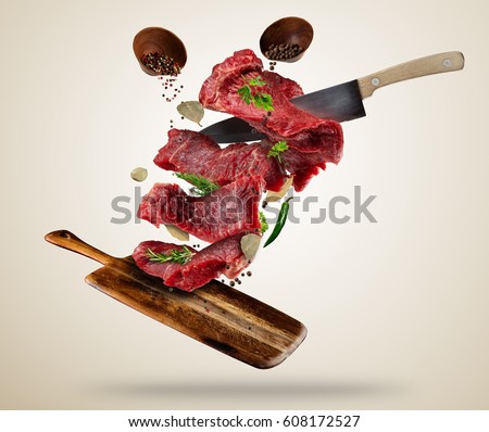 Flying pieces of raw steaks, with ingredients for cooking, served on wooden board. Knife cutting the meat. Concept of food preparation in low gravity mode. Separated on smooth background