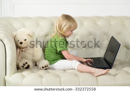 A little girl sits on the couch and plays with the computer (laptop). Sad toy Teddy bear sitting behind the girls. Photo about early child development and the dangers of computers for children