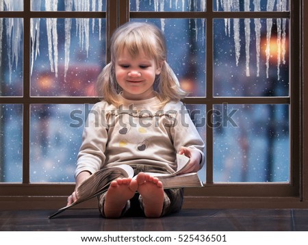 Little happy child reading a book. A large picture window. Outside the window, snow falls, winter. The house is warm, the girl barefoot