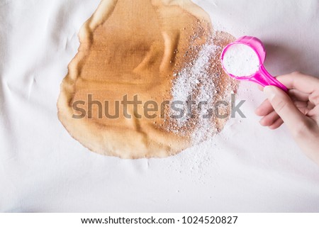 Big, brown coffee stain on light, white cloth. Woman\'s hand strewing washing powder from a cup or scoop. Spilled beverage stain removing. Dry cleaning concept. Clothes care. Chores of housewife.