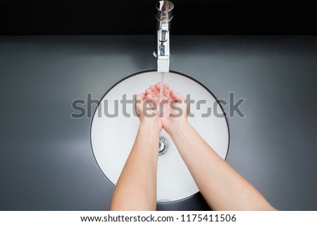 Young woman washing hands under the water tap. Water running. White, modern ceramic sink with dark gray table surface. Daily hygiene. Point of view shot. Contemporary restroom. Part of body.