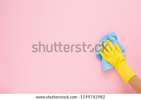 Employee hand in yellow rubber protective glove wiping pastel pink wall from dust with blue dry rag. General or regular cleanup. Commercial cleaning company. Copy space. Empty place for text or logo.