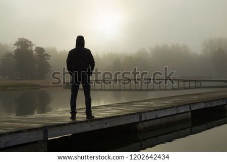 Young man standing alone on lake footbridge and staring at sunrise in gray, cloudy sky. Mist over water. Foggy air. Early chilly morning. Dark, scary moment and gloomy atmosphere. Back view.