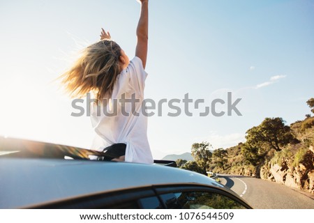 Beautiful and happy young woman, full of emotions and feelings enjoys warm summer air and sunshine on sunny day, stands up in car out of convertible cabriolet, wind in hair full of freedom