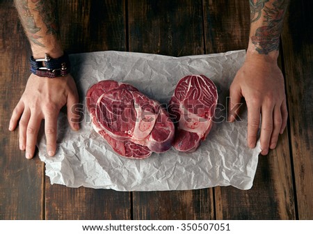 Top view of two tattooed hands and three steaks on white craft paper on old wooden table