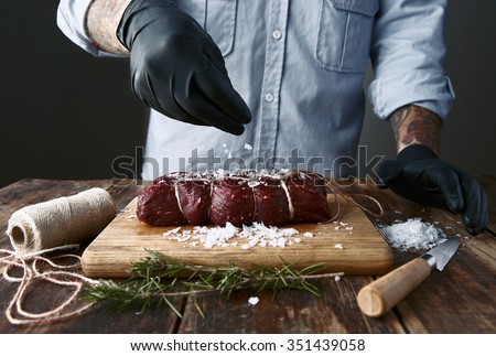 Tattoed butcher in black gloves salts tied piece of meat to smoke it.