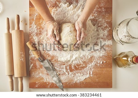 Top view on white table with isolated wooden board with knife, two rolling pins, bottle olive oil, transparent jar with flour Woman hands hold prepared dough for pasta or dumplings
