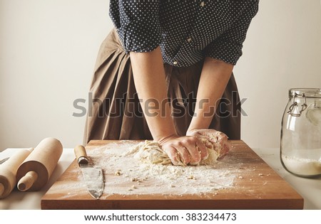 Woman kneads dough for pasta on wooden board near two rolling pins and jar with flour