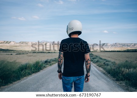 Photo from the back of brutal masculine man with authentic arm tattoos in blank black mockup tshirt standing in middle of desert highway