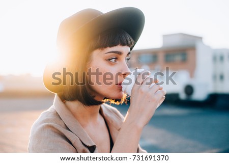 Attractive adorable woman with natural makeup sips on coffee in early morning on sunrise or sunset with light leaks, from to go cup, wears hipster fedora hat