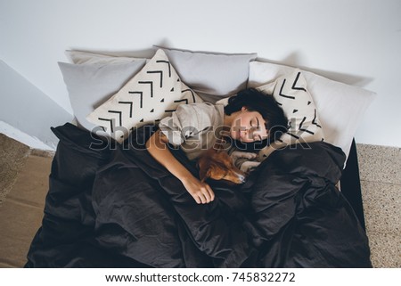 Young woman sleeps in bed, in clean and white simple bedroom, with her best friend, basenji breed puppy dog sleeping next to her, on top of covers. cute and adorable friendship