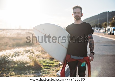 Portrait of young professional surfer man with tattoos and beard, who wears blank black tshirt and holds his surfboard under arm. Light leak from sunset sun behind shoulder. relaxed lifestyle