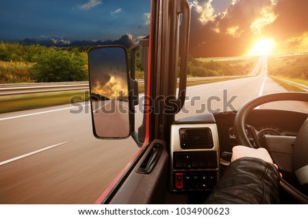 Truck dashboard with driver\'s hand on the steering wheel and side rear-view mirror on the countryside road against night sky with sunset