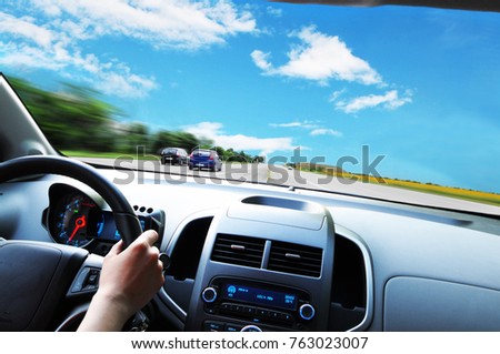 Car dashboard with driver's hand on the black steering wheel against asphalt road and blue sky with clouds