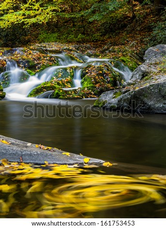 long exposure fall color with a waterfall, a swirling leaves in the foreground and lush greens with fall colors in the distance