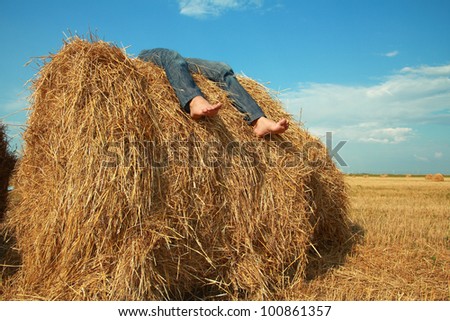 young barefoot girl lies on roll of dry hay under clear sky. Only feet are visible.